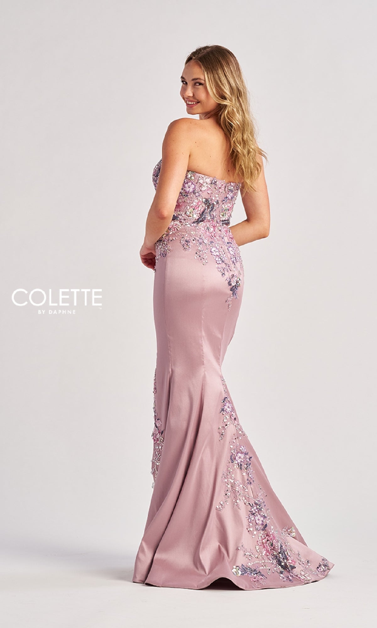 A woman in the Colette CL8410, a strapless light pink floor-length prom dress with floral embroidery, stands turned slightly away and smiles over her shoulder against a plain backdrop.